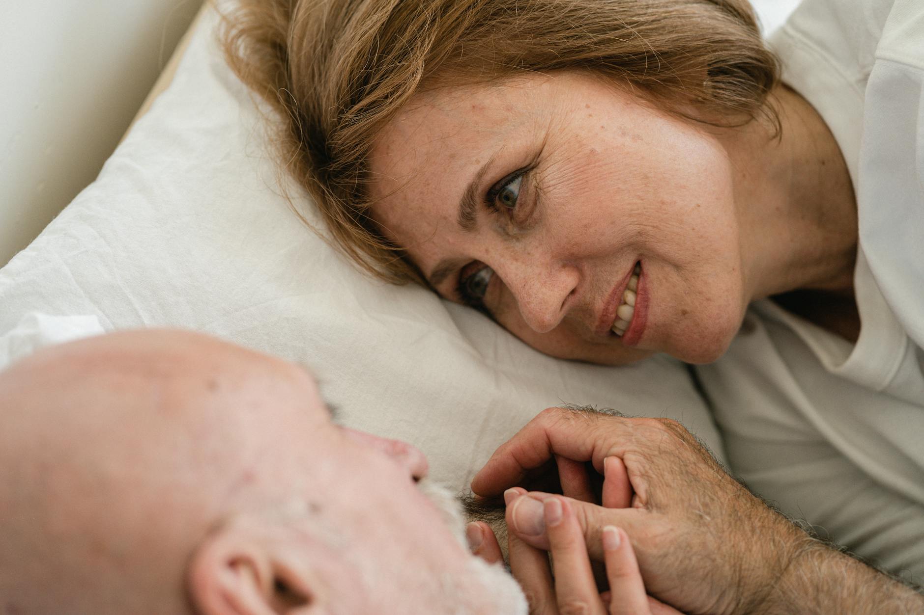 Starsze małżeństwo trzyma się za ręce w łóżku i patrzy sobie w oczy