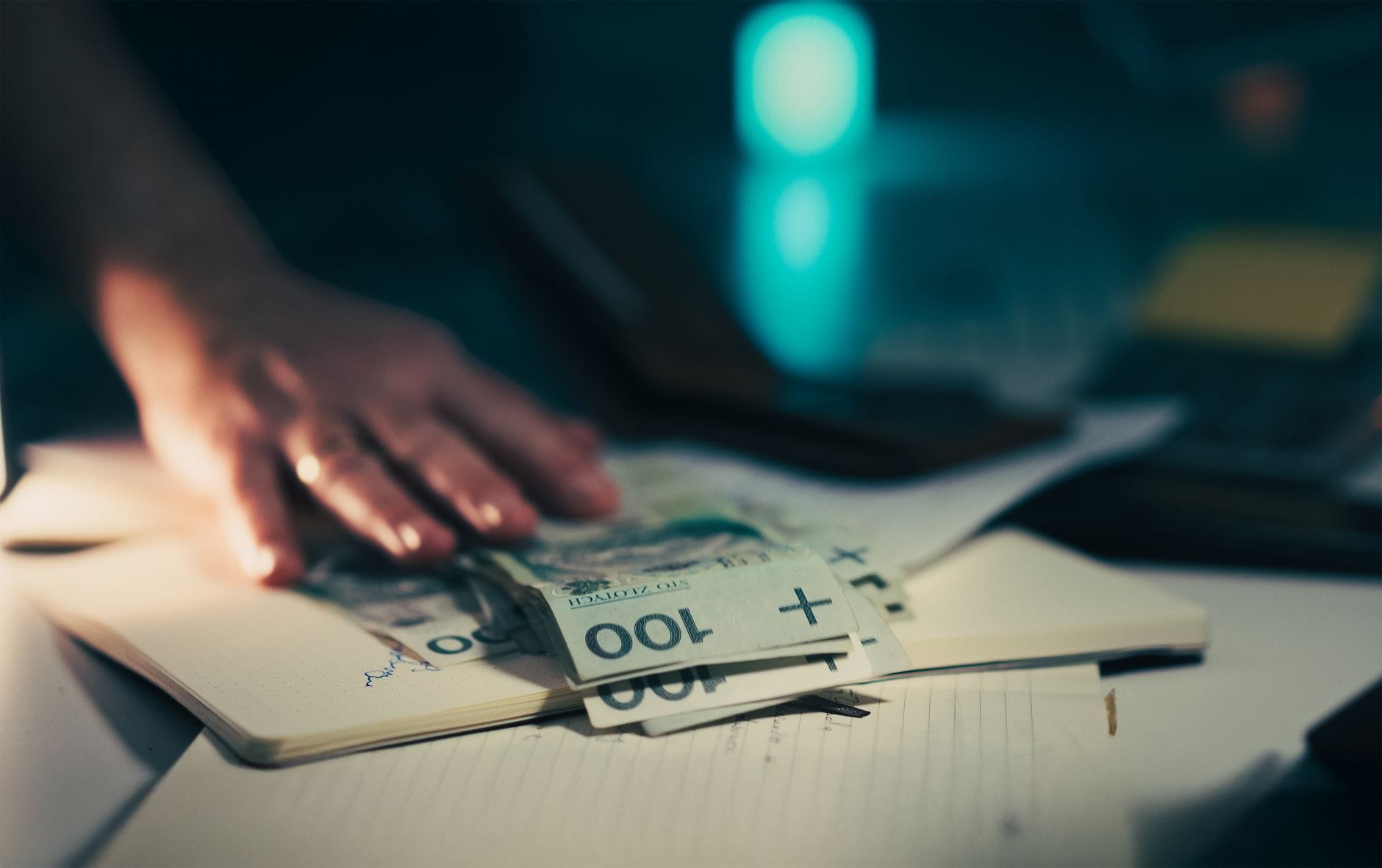 Dłoń sięga po banknoty leżące na biurku obok kalkulatora i notesu